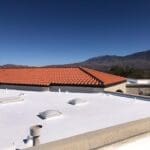 Flat white roof with multiple skylights and vents, adjacent to a building with a red tile roof, showing a well-maintained roofing surface under a clear blue sky with mountains in the background.