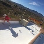 Workers applying a white coating to a flat roof with parapet walls, showing a maintenance process under a clear blue sky with desert landscape and mountains in the background.