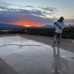 A landscape image of a Tucson home with a flat roof, freshly coated with a reflective or elastomeric roof coating. The setting includes desert landscaping with cacti, a bright sunny sky, and visible heat reflection from the roof.