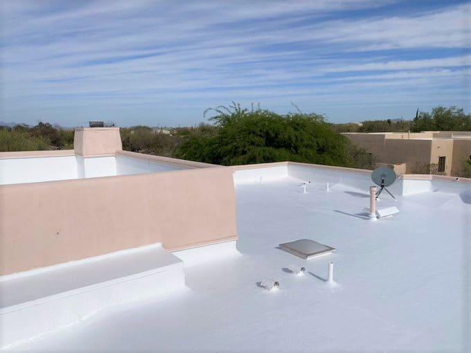 A landscape image of a Tucson home with a roof featuring a freshly applied reflective coating. The scene includes desert landscaping with cacti and succulents, a bright sunny sky, and an air conditioning unit visible near the house, emphasizing energy efficiency.